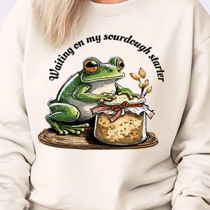 Pode incluir: Camisola creme com um desenho de um sapo segurando um pote de fermento natural. O sapo tem olhos grandes e laranja. O texto "Waiting on my sourdough starter" está acima. O sapo e o pote estão em madeira.