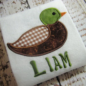 May include: A brown and green embroidered applique of a duck with a checkered pattern on its chest. The name "LIAM" is embroidered in green below the duck.