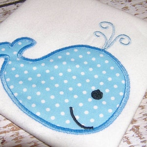 Puede incluir: Una ballena azul con lunares blancos y un ojo negro, bordada en una tela blanca.