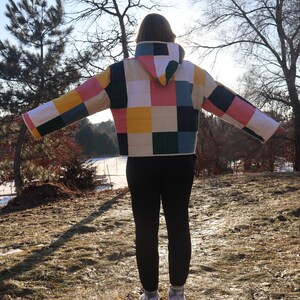 May include: A quilted jacket with a patchwork design in various colors, including pink, yellow, green, and blue. The jacket has a hood and is worn with black leggings and white sneakers. The person is standing outdoors in a natural setting.