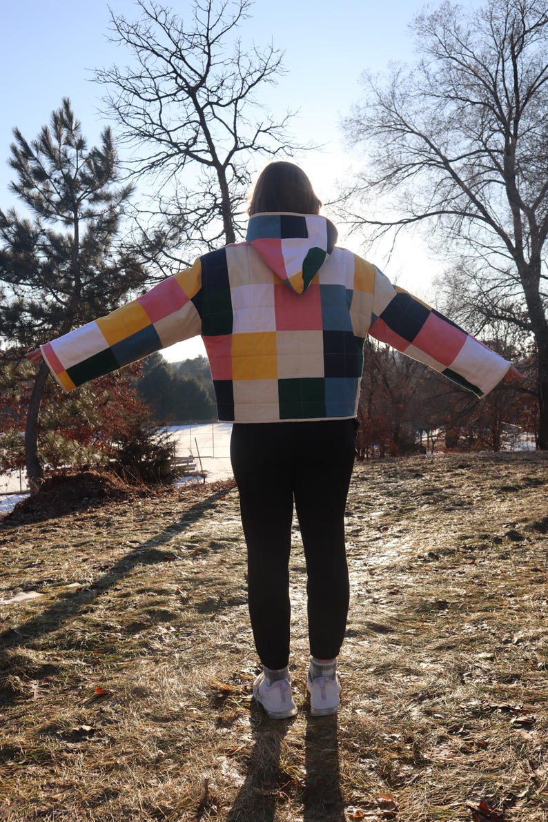 May include: A quilted jacket with a patchwork design in various colors, including pink, yellow, green, and blue. The jacket has a hood and is worn with black leggings and white sneakers. The person is standing outdoors in a natural setting.