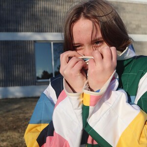 May include: A colorful quilted jacket with a patchwork design. The jacket features a variety of colors, including pink, blue, yellow, and green. The sleeves have white cuffs. The person is holding a white object near their face.