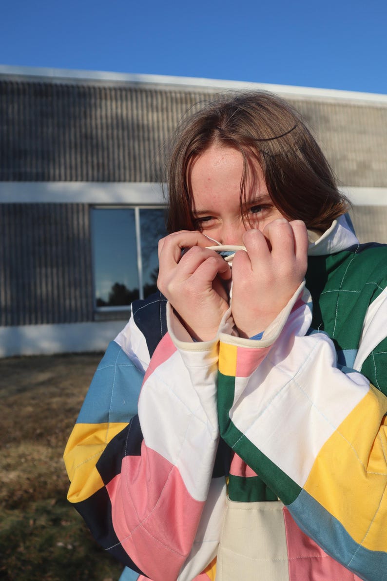 May include: A colorful quilted jacket with a patchwork design. The jacket features a variety of colors, including pink, blue, yellow, and green. The sleeves have white cuffs. The person is holding a white object near their face.