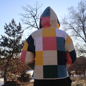 May include: A quilted jacket with a colorful patchwork design. The jacket features a hood and a square pattern in shades of pink, yellow, green, blue, and cream. The person is wearing black pants. The background includes trees and a clear sky.