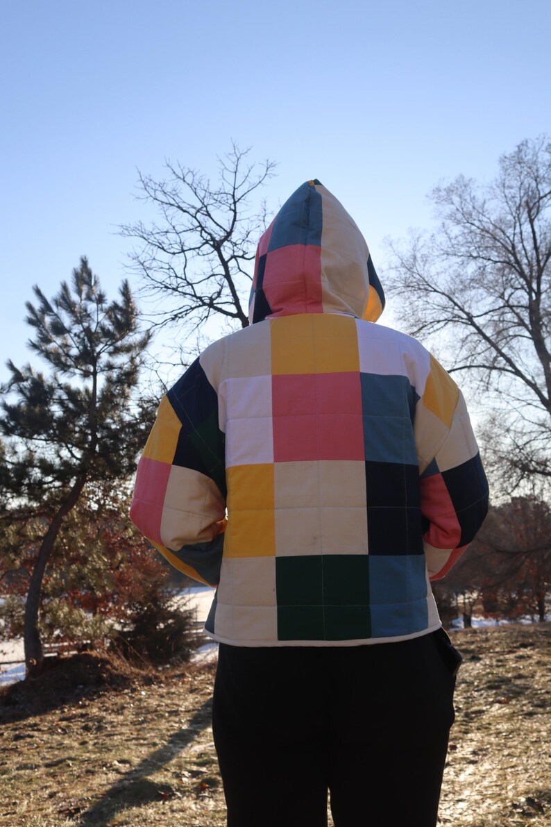 May include: A quilted jacket with a colorful patchwork design. The jacket features a hood and a square pattern in shades of pink, yellow, green, blue, and cream. The person is wearing black pants. The background includes trees and a clear sky.