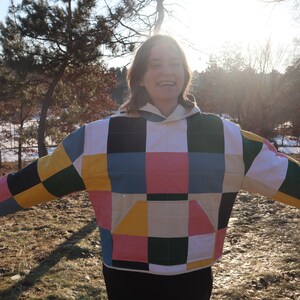May include: A colorful patchwork jacket with a hood. The jacket features a pattern of squares in various colors, including yellow, pink, blue, green, and black. The jacket is worn outdoors in a natural setting.