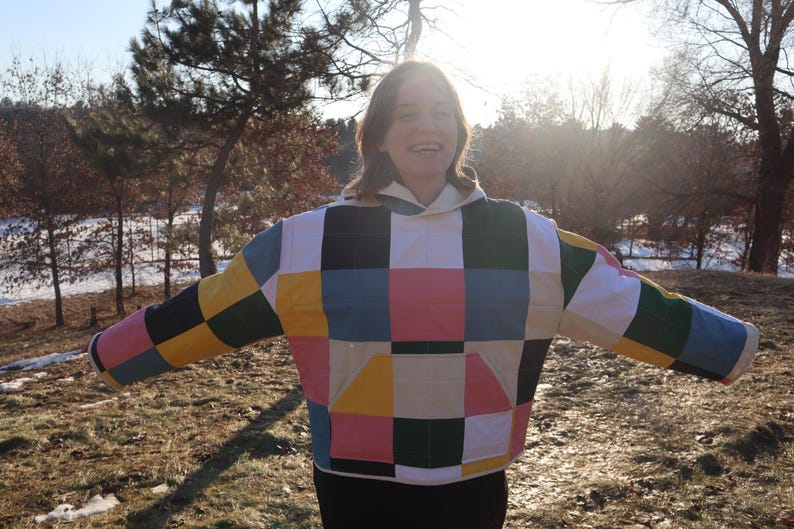 May include: A colorful patchwork jacket with a hood. The jacket features a pattern of squares in various colors, including yellow, pink, blue, green, and black. The jacket is worn outdoors in a natural setting.