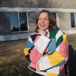 May include: A colorful quilted jacket with a patchwork design. The jacket features squares of pink, white, yellow, blue, and green. The jacket has a white collar and cuffs. The person is wearing black pants.