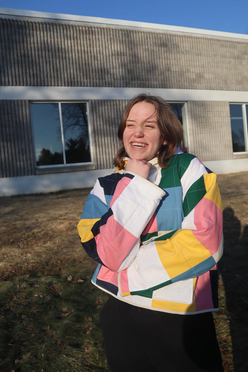 May include: A colorful quilted jacket with a patchwork design. The jacket features squares of pink, white, yellow, blue, and green. The jacket has a white collar and cuffs. The person is wearing black pants.