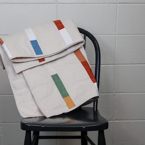 May include: A folded beige quilt with colorful rectangular accents in red, white, blue, green, and orange rests on a black wooden chair. The quilt's design features a minimalist, geometric pattern.