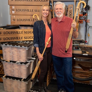 Puede incluir: Dos bastones de madera de color claro, cada uno con un mango curvo y el logotipo de Louisville Slugger, son sostenidos por dos personas. Los bastones tienen un acabado liso y pulido con una punta de goma negra en la base.
