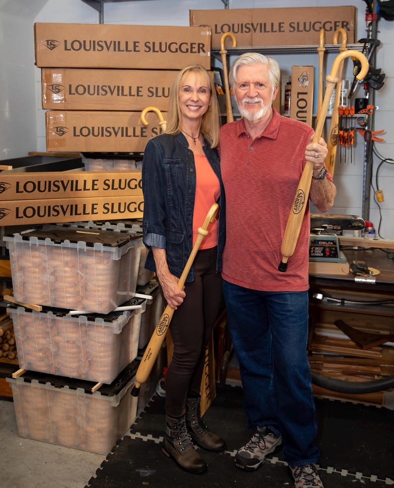 Puede incluir: Dos bastones de madera de color claro, cada uno con un mango curvo y el logotipo de Louisville Slugger, son sostenidos por dos personas. Los bastones tienen un acabado liso y pulido con una punta de goma negra en la base.