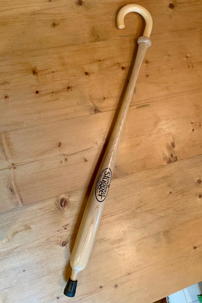 Puede incluir: Un bast&oacute;n de madera de color claro fabricado a partir de un bate de b&eacute;isbol, con un mango curvado en la parte superior y una punta de goma negra en la base. El cuerpo muestra el logotipo negro de Louisville Slugger impreso en la madera.