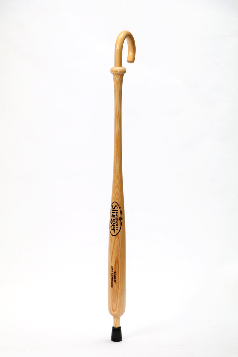 Puede incluir: Un bast&oacute;n de madera de color claro fabricado a partir de un bate de b&eacute;isbol, con un mango curvado en la parte superior y una punta de goma negra en la base. El cuerpo muestra el texto impreso en negro Louisville Slugger y Slugger John Camistrare.
