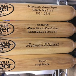 Puede incluir: Cuatro bates de b&eacute;isbol de madera clara con el logotipo negro de Louisville Slugger y texto grabado con l&aacute;ser. Los bates muestran nombres y fechas, incluyendo Southwest Lamar Tigers, Coach Jay Stahl, Mason Max, Maria's Papa, Herman Stewart y Virgil Mione.