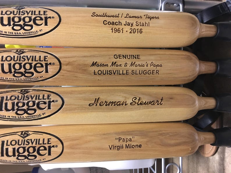 Puede incluir: Cuatro bates de b&eacute;isbol de madera clara con el logotipo negro de Louisville Slugger y texto grabado con l&aacute;ser. Los bates muestran nombres y fechas, incluyendo Southwest Lamar Tigers, Coach Jay Stahl, Mason Max, Maria's Papa, Herman Stewart y Virgil Mione.