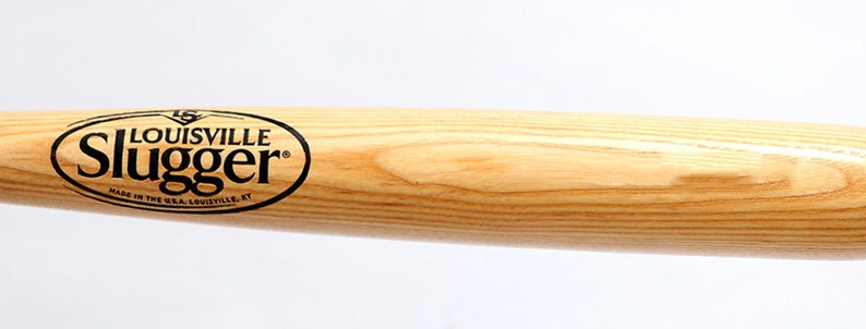 Puede incluir: Un bate de b&eacute;isbol de madera clara con un logotipo ovalado negro con el texto Louisville Slugger y Made in the USA, Louisville, KY impreso en el lateral. La veta de la madera es visible en toda la superficie del bate cil&iacute;ndrico.