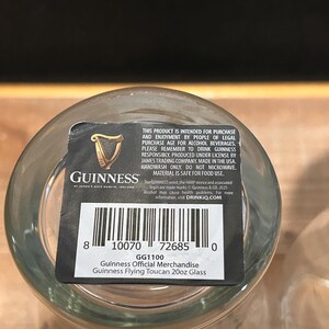 May include: A clear Guinness Flying Toucan 20oz glass with a black and white label. The label includes the Guinness logo, a harp, and the text "Guinness Official Merchandise". The glass is made in the USA.