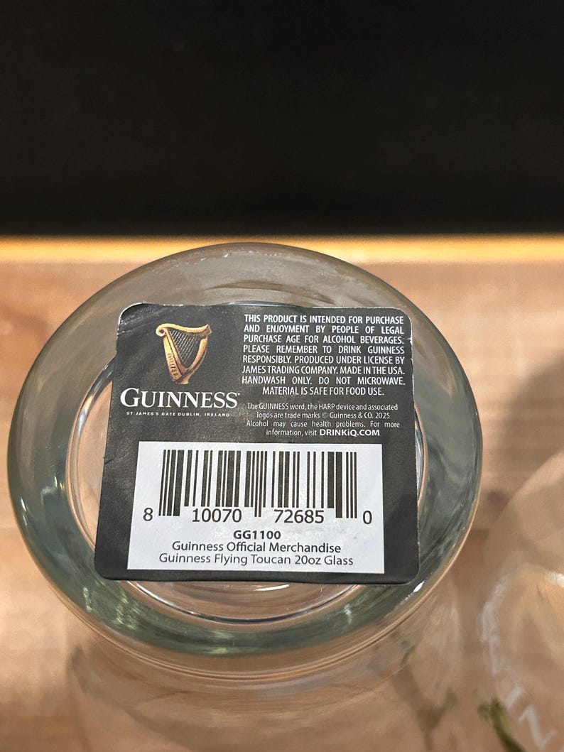 May include: A clear Guinness Flying Toucan 20oz glass with a black and white label. The label includes the Guinness logo, a harp, and the text "Guinness Official Merchandise". The glass is made in the USA.