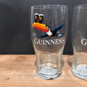 May include: Two clear Guinness pint glasses. One glass features a colorful toucan graphic with two pints of Guinness and the Guinness logo. The glasses are on a wooden surface.