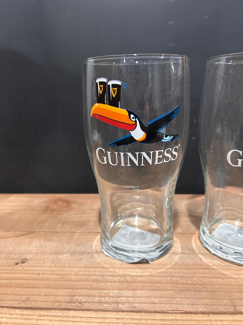 May include: Two clear Guinness pint glasses. One glass features a colorful toucan graphic with two pints of Guinness and the Guinness logo. The glasses are on a wooden surface.