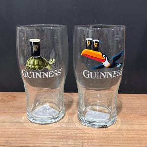 May include: Two clear Guinness pint glasses. One glass features a turtle with a Guinness pint, the other a toucan with two Guinness pints. The Guinness logo is printed on both glasses.