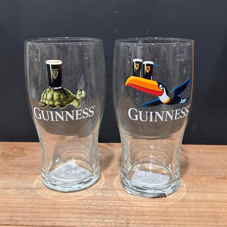 May include: Two clear Guinness pint glasses. One glass features a turtle with a Guinness pint, the other a toucan with two Guinness pints. The Guinness logo is printed on both glasses.