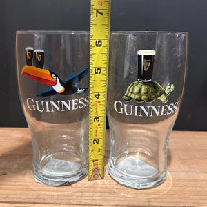 May include: Two clear Guinness pint glasses. One glass features a toucan with a Guinness pint, and the other has a turtle with a Guinness pint. The glasses are approximately 6 inches tall.