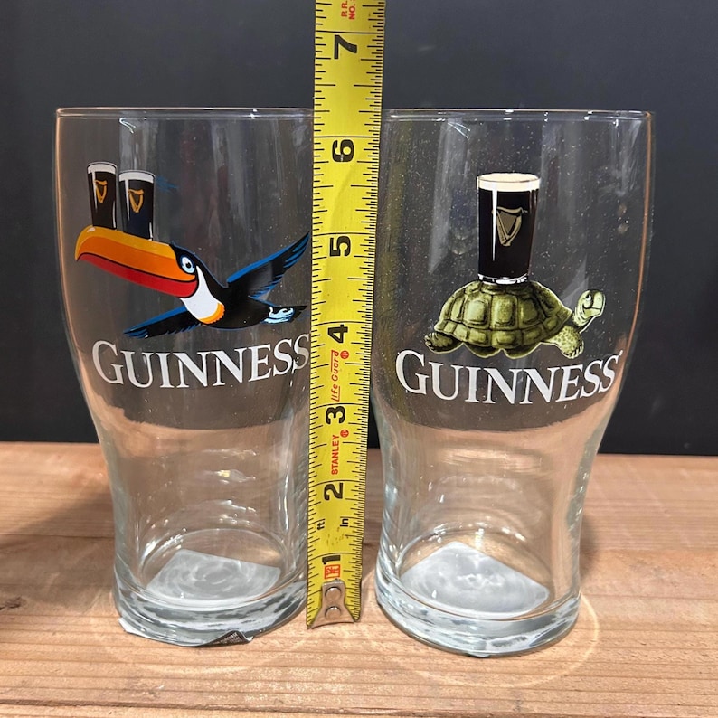 May include: Two clear Guinness pint glasses. One glass features a toucan with a Guinness pint, and the other has a turtle with a Guinness pint. The glasses are approximately 6 inches tall.