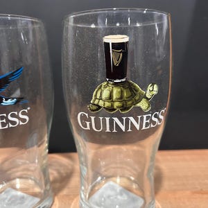 May include: Clear pint glass with a Guinness logo featuring a turtle with a pint of Guinness on its back. The word "GUINNESS" is printed in white on the glass. The glass is empty and has a triangular base.