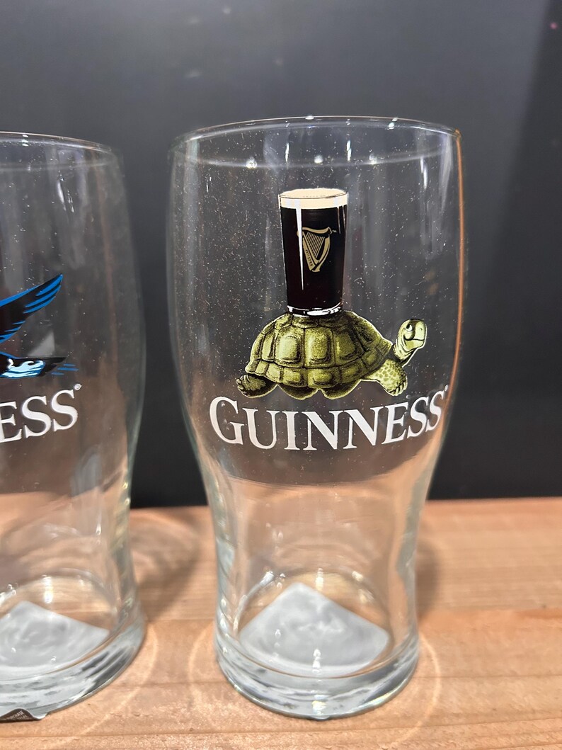 May include: Clear pint glass with a Guinness logo featuring a turtle with a pint of Guinness on its back. The word "GUINNESS" is printed in white on the glass. The glass is empty and has a triangular base.