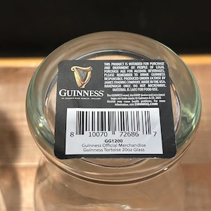 May include: A clear Guinness Official Merchandise Tortoise 20oz glass. The glass has a black and white label with the Guinness logo, a harp, and product information. The label also includes a barcode and the text "GG1200".