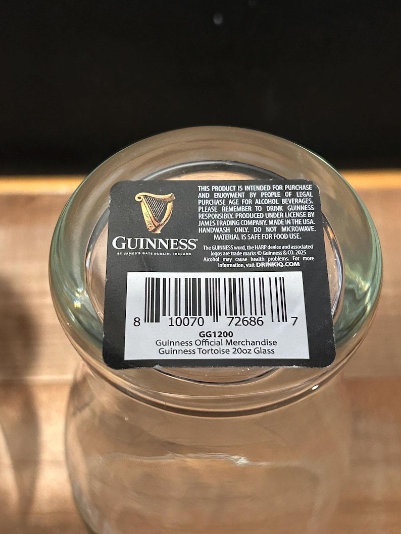May include: A clear Guinness Official Merchandise Tortoise 20oz glass. The glass has a black and white label with the Guinness logo, a harp, and product information. The label also includes a barcode and the text "GG1200".