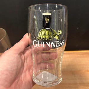 May include: A clear pint glass with the Guinness logo and a whimsical design of a turtle with a Guinness pint glass on its back. The word "Guinness" is printed in white on the glass.