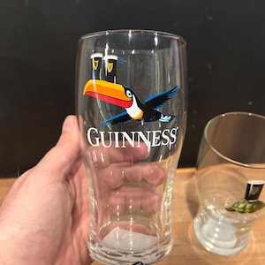 May include: Clear pint glass with the Guinness logo and a toucan bird graphic. The toucan has a large orange and yellow beak, with black and blue wings. The word "GUINNESS" is printed in white.