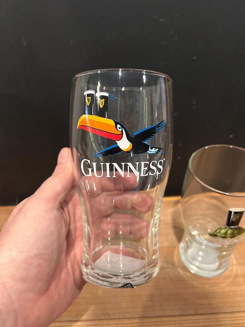 May include: Clear pint glass with the Guinness logo and a toucan bird graphic. The toucan has a large orange and yellow beak, with black and blue wings. The word "GUINNESS" is printed in white.