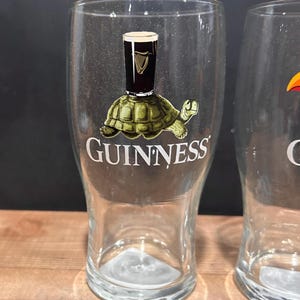 May include: Clear pint glass with a Guinness logo. The logo features a turtle carrying a pint of Guinness. The word "GUINNESS" is printed below the turtle. The glass is on a wooden surface.