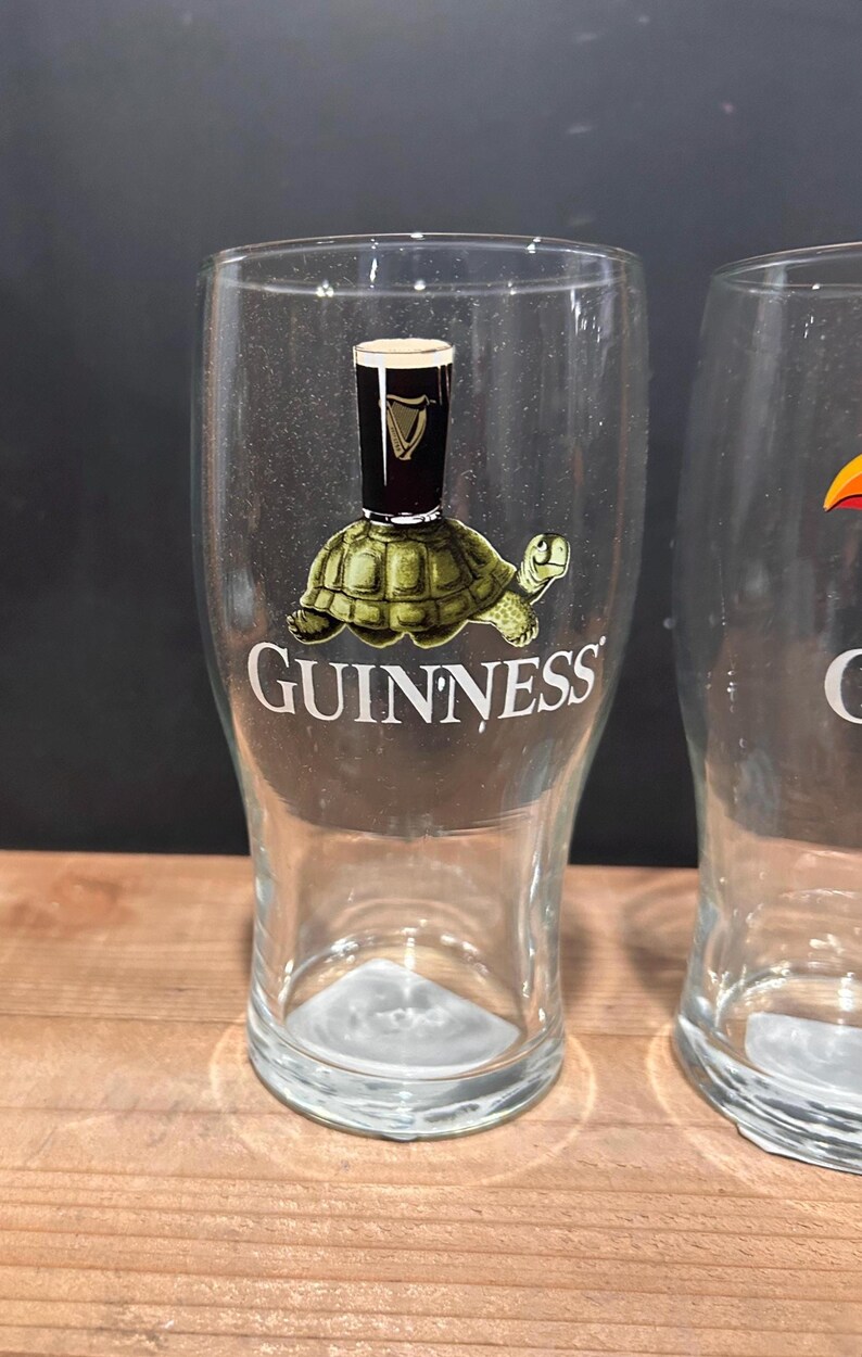May include: Clear pint glass with a Guinness logo. The logo features a turtle carrying a pint of Guinness. The word "GUINNESS" is printed below the turtle. The glass is on a wooden surface.
