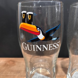 May include: Clear pint glass with a toucan design and the word "GUINNESS" in white. The toucan has a black body, blue wings, and a large orange and red beak. Two pints of stout are above the toucan.