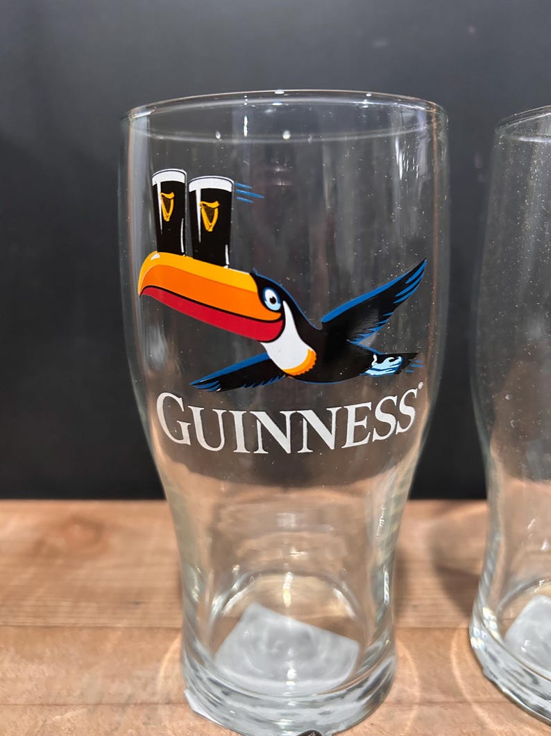 May include: Clear pint glass with a toucan design and the word "GUINNESS" in white. The toucan has a black body, blue wings, and a large orange and red beak. Two pints of stout are above the toucan.