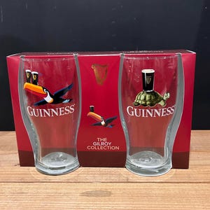 May include: Two clear Guinness pint glasses with colorful toucan and turtle designs. The glasses are in a red box with the Guinness logo and "The Gilroy Collection" text.