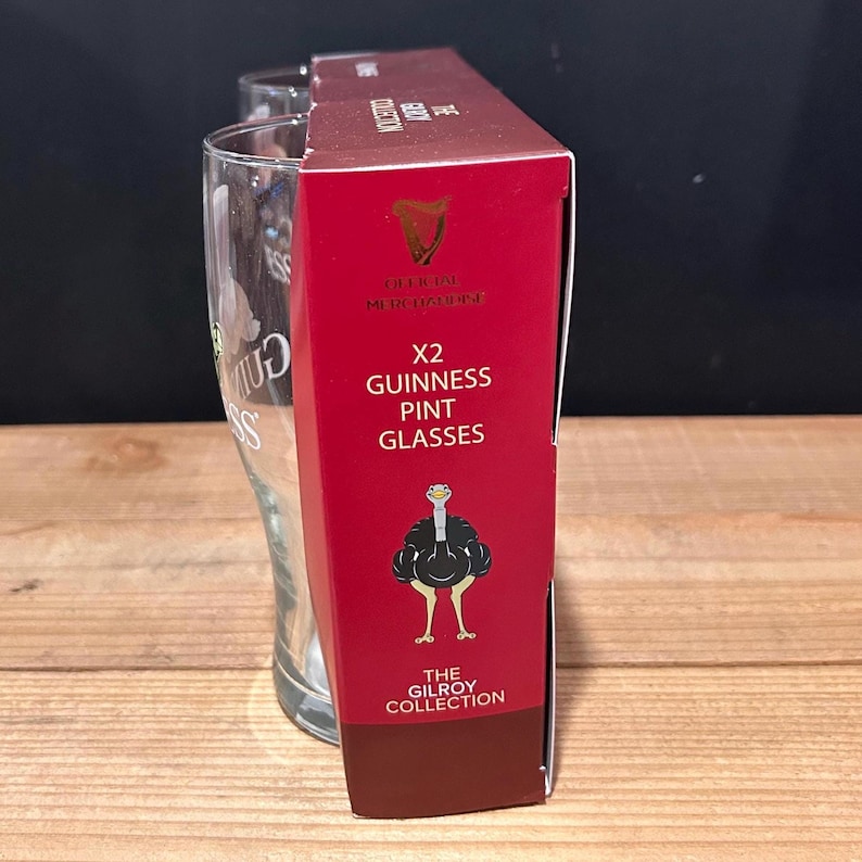 May include: A red box with the text "X2 Guinness Pint Glasses" and an illustration of an ostrich. Two pint glasses are visible behind the box. The box also has the text "The Gilroy Collection" and the Guinness logo.