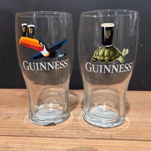 May include: Two clear Guinness pint glasses. One glass features a toucan with a Guinness pint, and the other has a turtle with a Guinness pint. The word "Guinness" is printed in white on both glasses.
