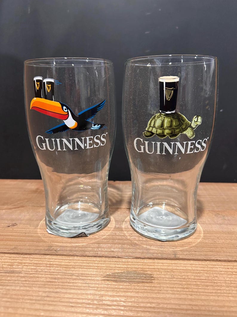 May include: Two clear Guinness pint glasses. One glass features a toucan with a Guinness pint, and the other has a turtle with a Guinness pint. The word "Guinness" is printed in white on both glasses.