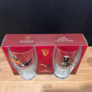 May include: A set of two Guinness glasses with a red box. The glasses feature toucan and turtle designs with the Guinness logo. The box reads "The Gilroy Collection" and displays the Guinness logo.