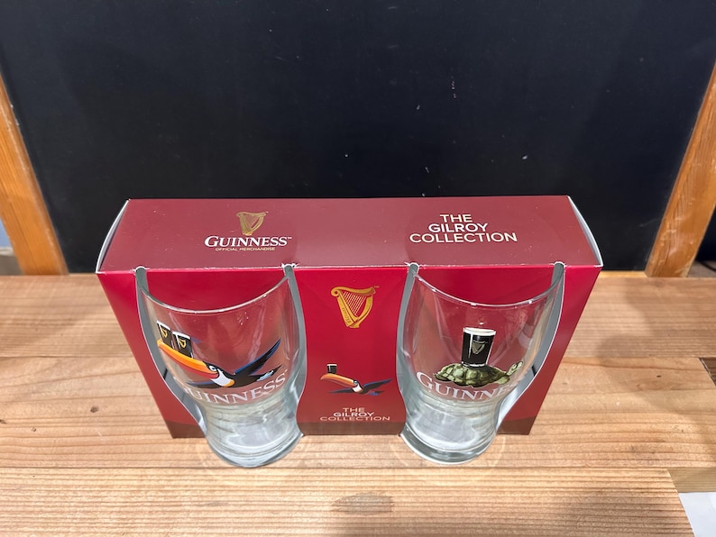 May include: A set of two Guinness glasses with a red box. The glasses feature toucan and turtle designs with the Guinness logo. The box reads "The Gilroy Collection" and displays the Guinness logo.