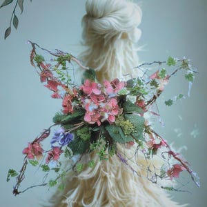 May include: An artistic composition featuring a central figure with a fluffy, cream-coloured texture, adorned with a floral arrangement of pink, purple, and green foliage. The image has a soft, dreamy aesthetic with the text "Florist Rhi".