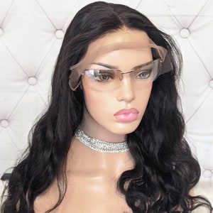 Może przedstawiać: Czarna peruka z włosów ludzkich z luźnymi falami. Peruka jest na głowie manekina i ma koronkę z przodu. Peruka jest ułożona z przedziałkiem na środku i ma naturalnie wyglądającą linię włosów.