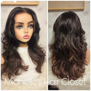 Może przedstawiać: Długa, ciemnobrązowa peruka z luźnymi falami. Peruka jest ułożona z przedziałkiem na środku i ma naturalnie wyglądającą linię włosów. Peruka jest pokazana na głowie manekina.