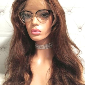 Może przedstawiać: Długa, falująca, brązowa peruka z ludzkich włosów na głowie manekina. Peruka jest ułożona w luźne fale i sięga do ramion. Manekin nosi przezroczyste okulary w czarnych oprawkach i srebrny choker z kryształkami.